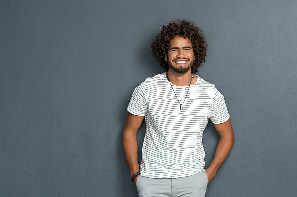 Portrait of happy young man with curly hair standing against grey background. African man with hands in pocket leaning against grey wall. Multi ethnic young man in casual looking at camera with copy space.